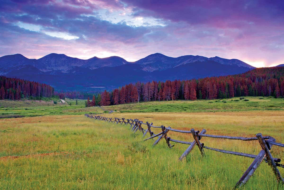 A wooden fence cuts across a green meadow as the setting sun changes the sky and mountain peaks purple