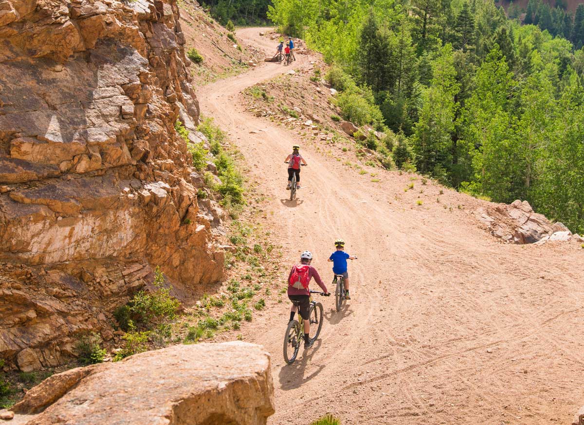 Three bikers ride along a gravel trail surrounded with green trees on one side and a rock wall on the other
