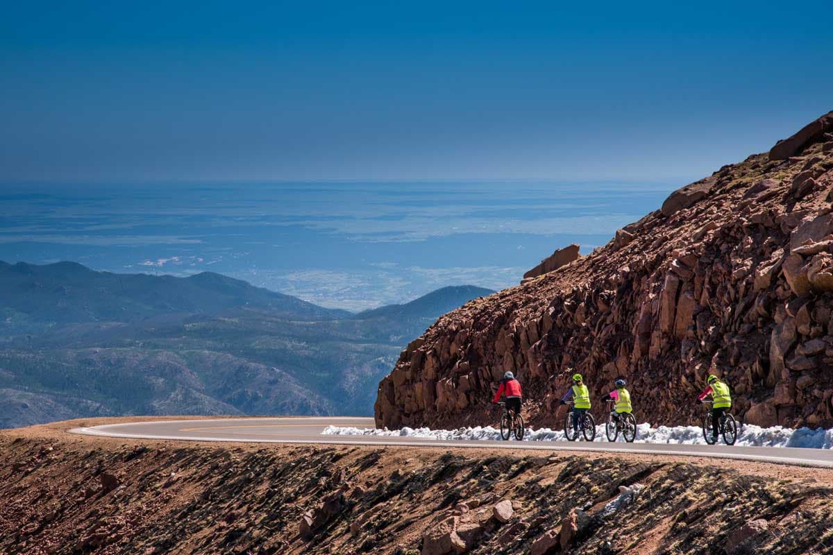 Four bikers in bright colors ride down Pikes Peak with mountains and a blue sky in the background