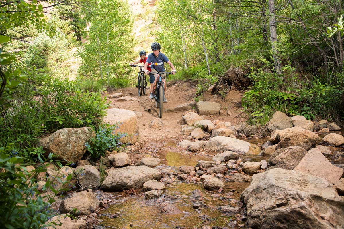 Two children on bikes cross a trickling creek surrounded by small rocks