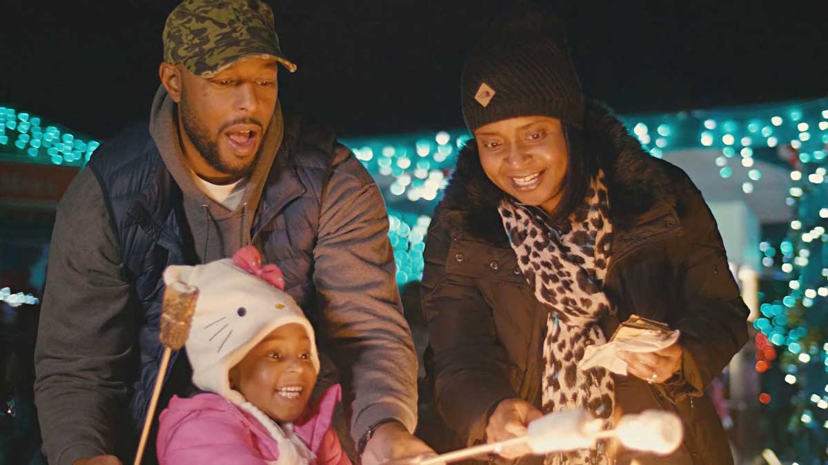 Two parents and a child roast marshmallows around a fire at the Denver Zoo in Colorado. In the background, blue holiday lights shine brightly.