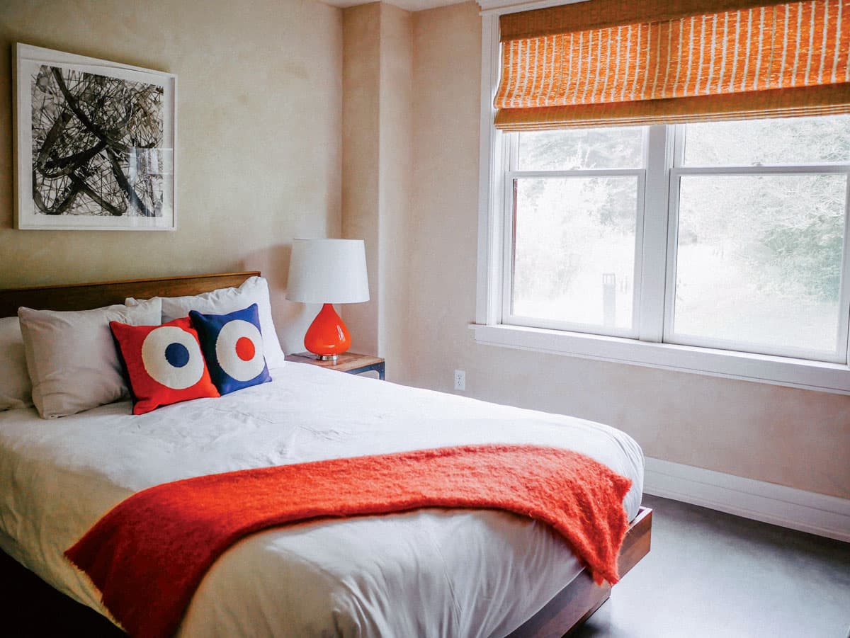 A bed with white linens surrounded by blue and orange accents with sun streaming in