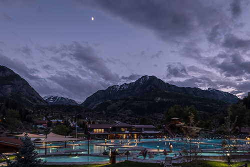 The light-blue pools of Ouray Hot Springs Pool sit under the night sky with clouds and half of the moon. There are snow-capped mountains in the distance.