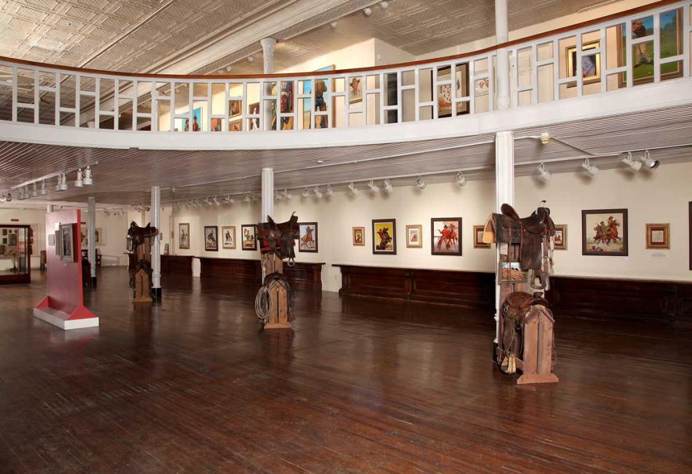 Saddles and riding accessories, like ropes and stirrups, are displayed on special pedestals throughout the first floor of an art museum in Trinidad, Colorado. Two floors of white walls are lined with outdoor-themed paitings. 