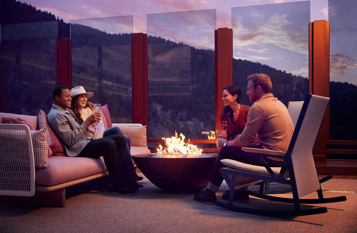 Two couples warm themselves around a fire while holding cocktails on a rooftop deck; a forest of evergreens can be seen in the distance