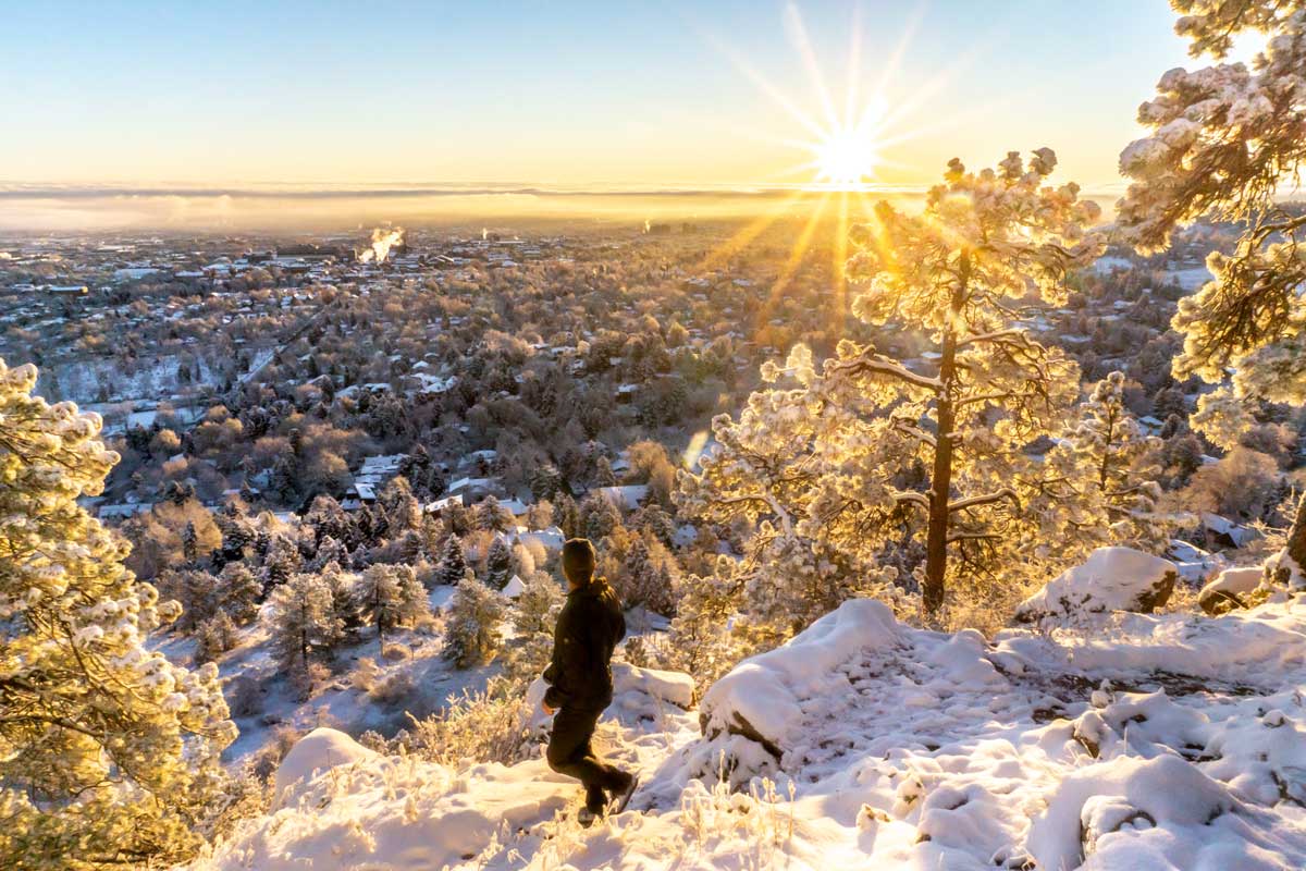 A hiker dressed in long black clothes and gray gloves makes their way down a snow-covered path in a wood in Colorado as the sunrises.
