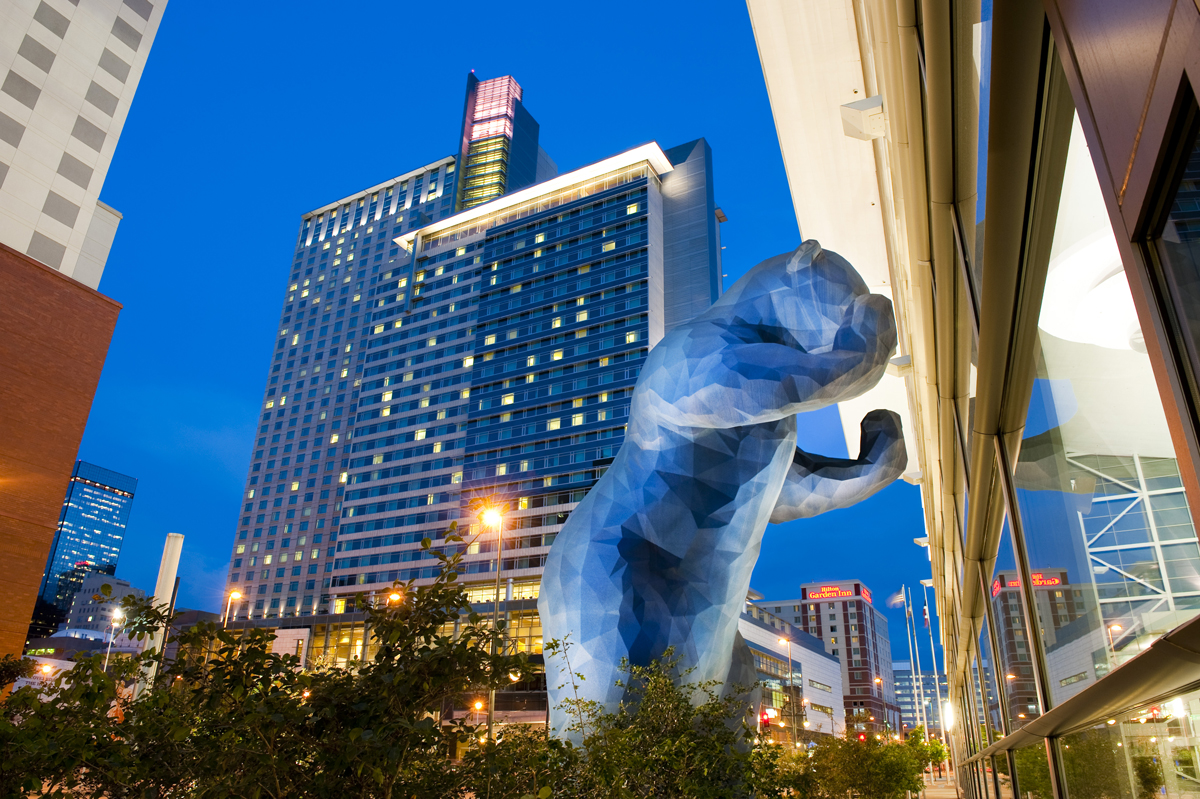 A photo looking up from feet away at the Big Blue Bear peering into the Convention Center in downtown Denver. There is a large hotel building behind the bear, underneath a dark-blue evening sky. 