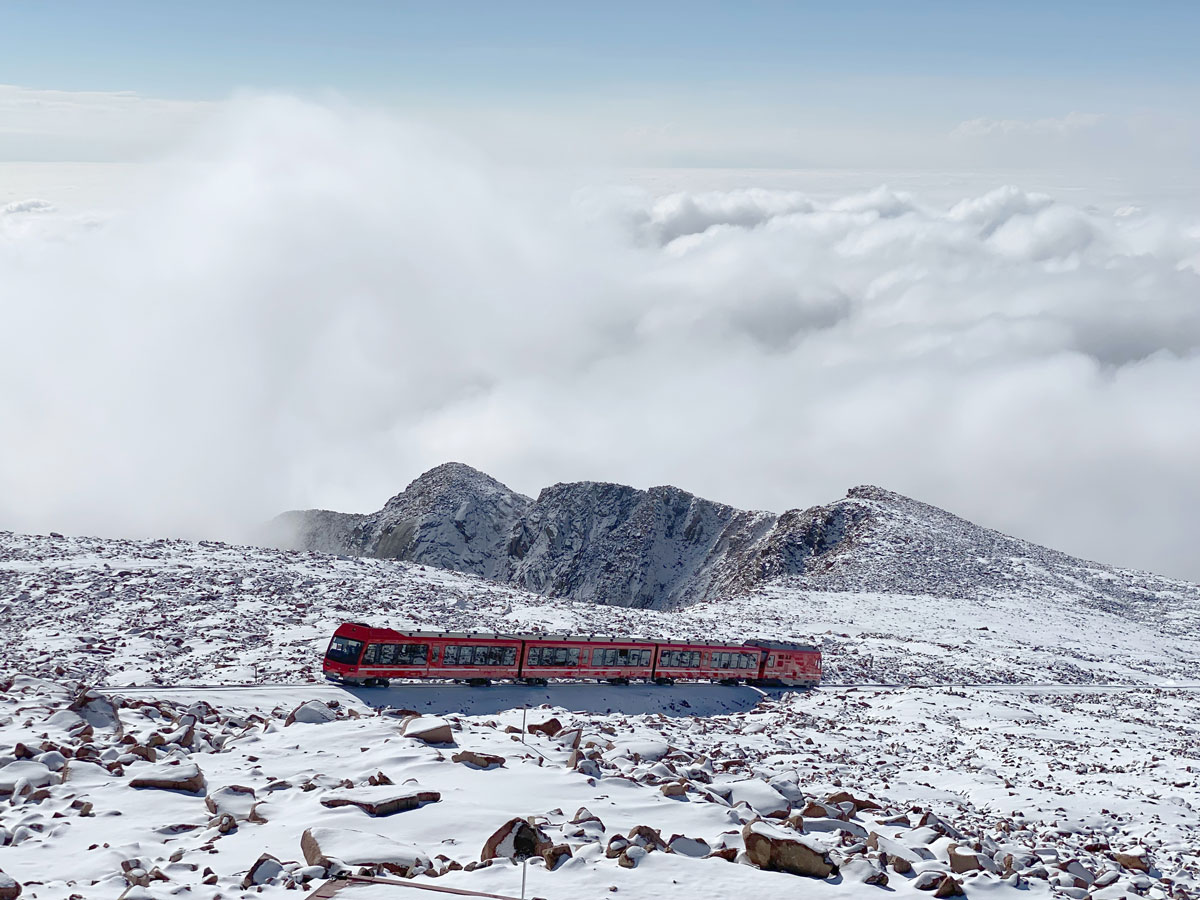 A short 4-piece train travels amid a snow-covered landscape. The train appears to be atop the world with no trees and a field of thick, fluffy clouds below the edge of the Colorado mountaintop.