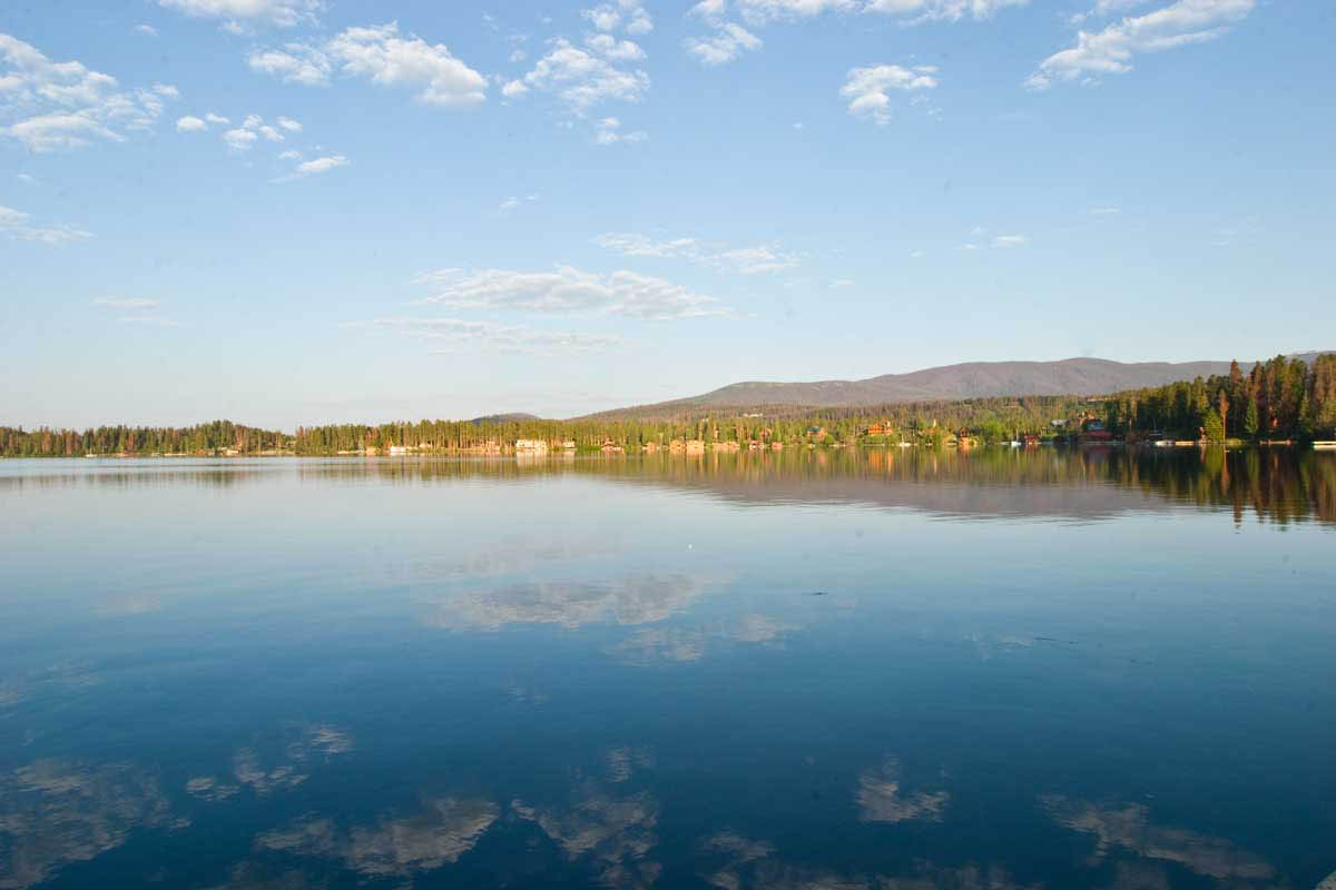 The waters of Grand Lake are still and calm. The lake reflects the blue sky dotted with white clouds above.