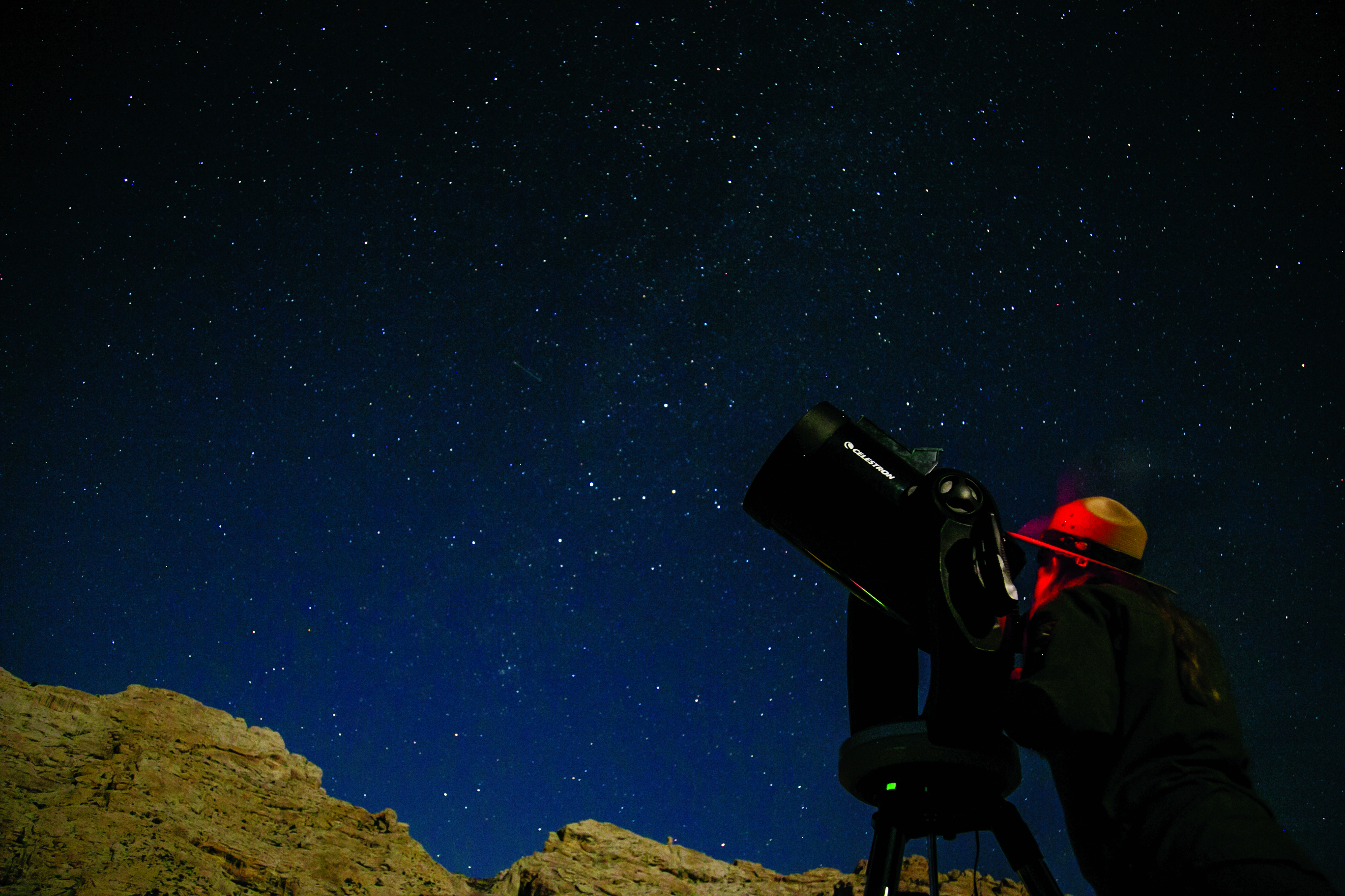 Someone in a hat looking into a telescope that's pointed at a star-studded sky.