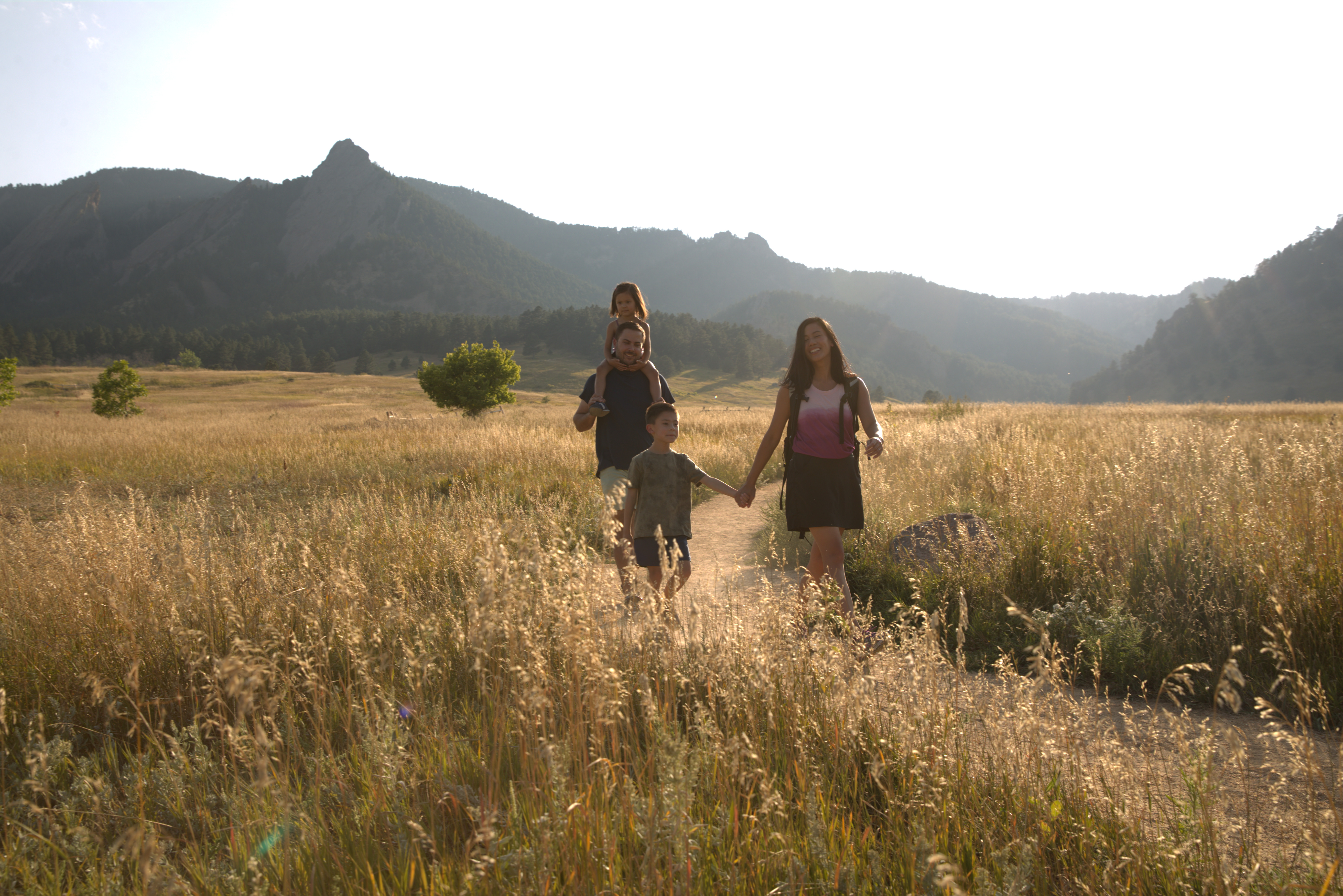 Two adults and a child hiking along a Chautauqua trail at dusk. The meadow surrounding them is almost as tall as the kid. We see the navy blue foothill mountanis behind them