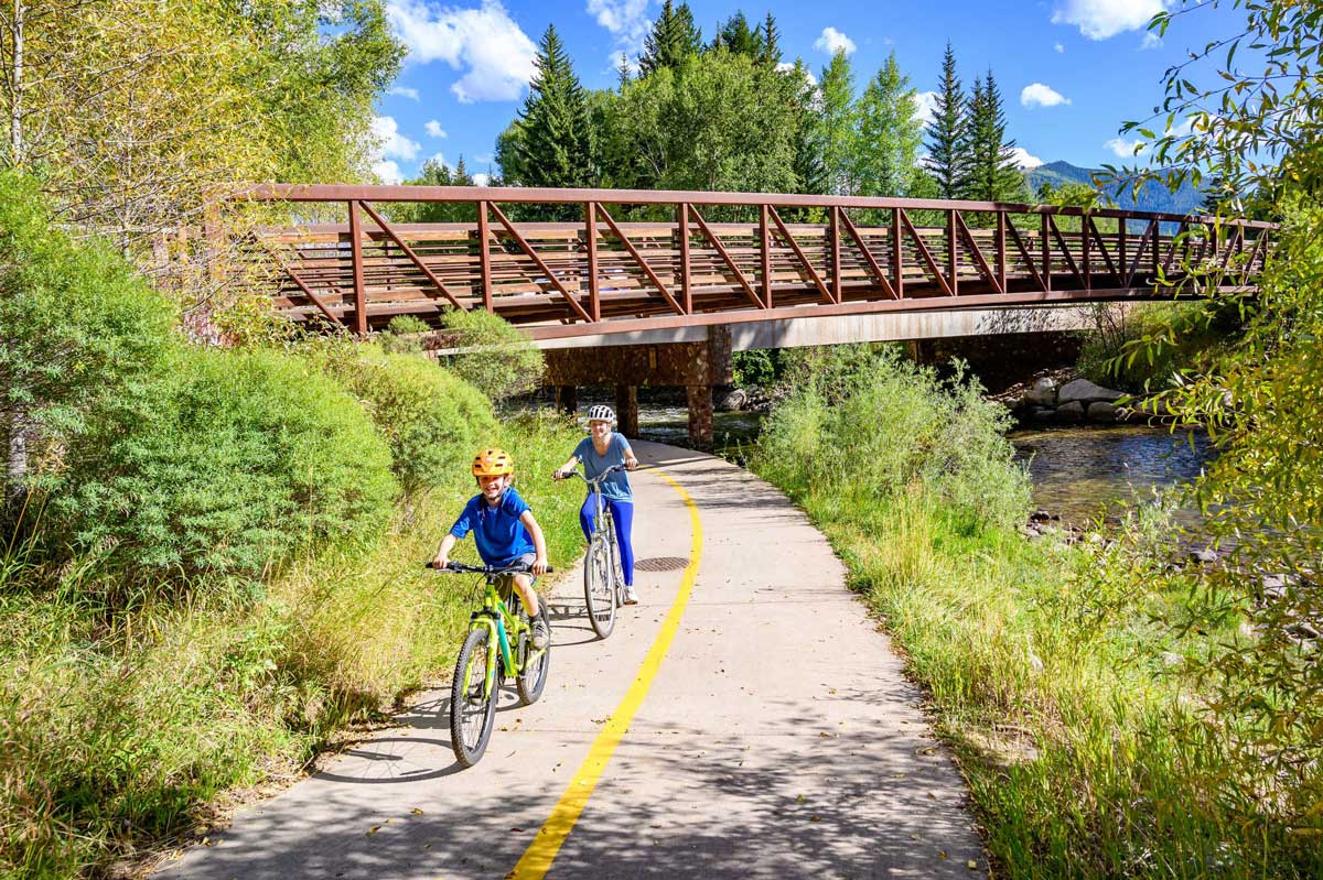 Kids biking on a paved path near a river with a bridge in the background. They're surrounded by green trees and shrubs.