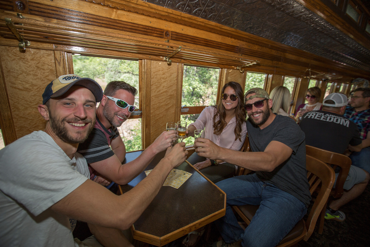 A group of four people clink beer glasses filled with amber suds as they sit in a historical train car with polished wooden walls and ornate carvings in Colorado.