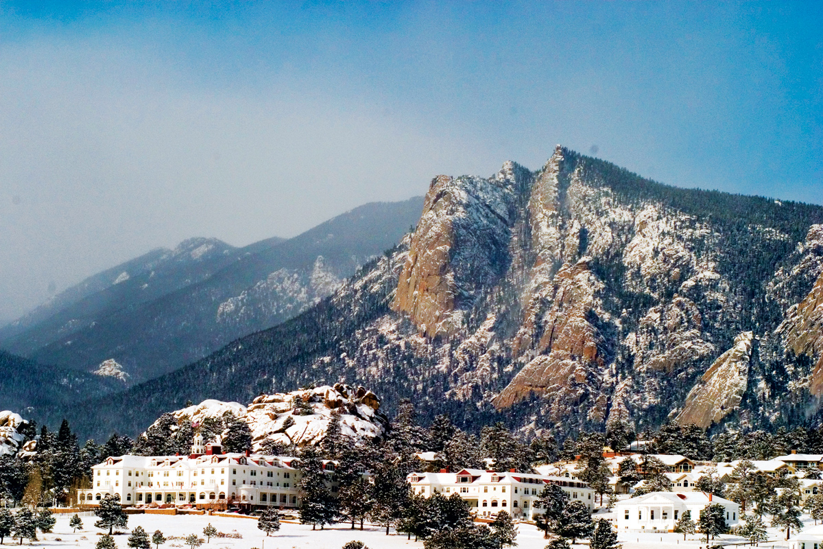 The red-roofed buildings of The Stanley Hotel in Estes Park are covered in a blanket of snow. A tall, cleaved mountain peak looms just being the hotel and is snow-covered.