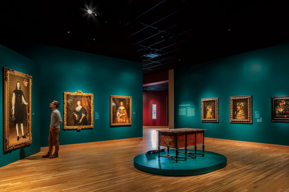 A large, jewel-tone blue-green room at the Denver Art Museum features a desk on a platform in the middle and walls filled with ornate portrait of people.