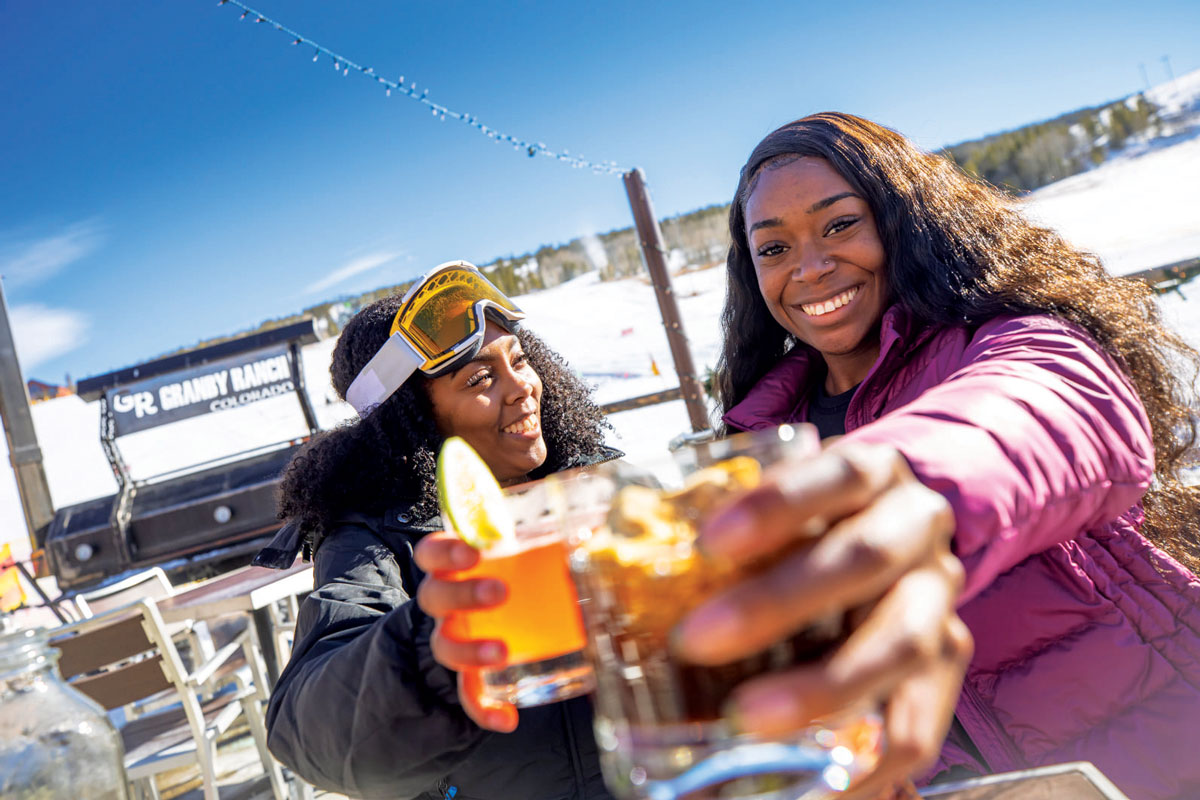 Two people smile and clink glasses of apres-ski cocktails at Ski Granby Ranch in Colorado. Behind them the landscape is covered in white snow.