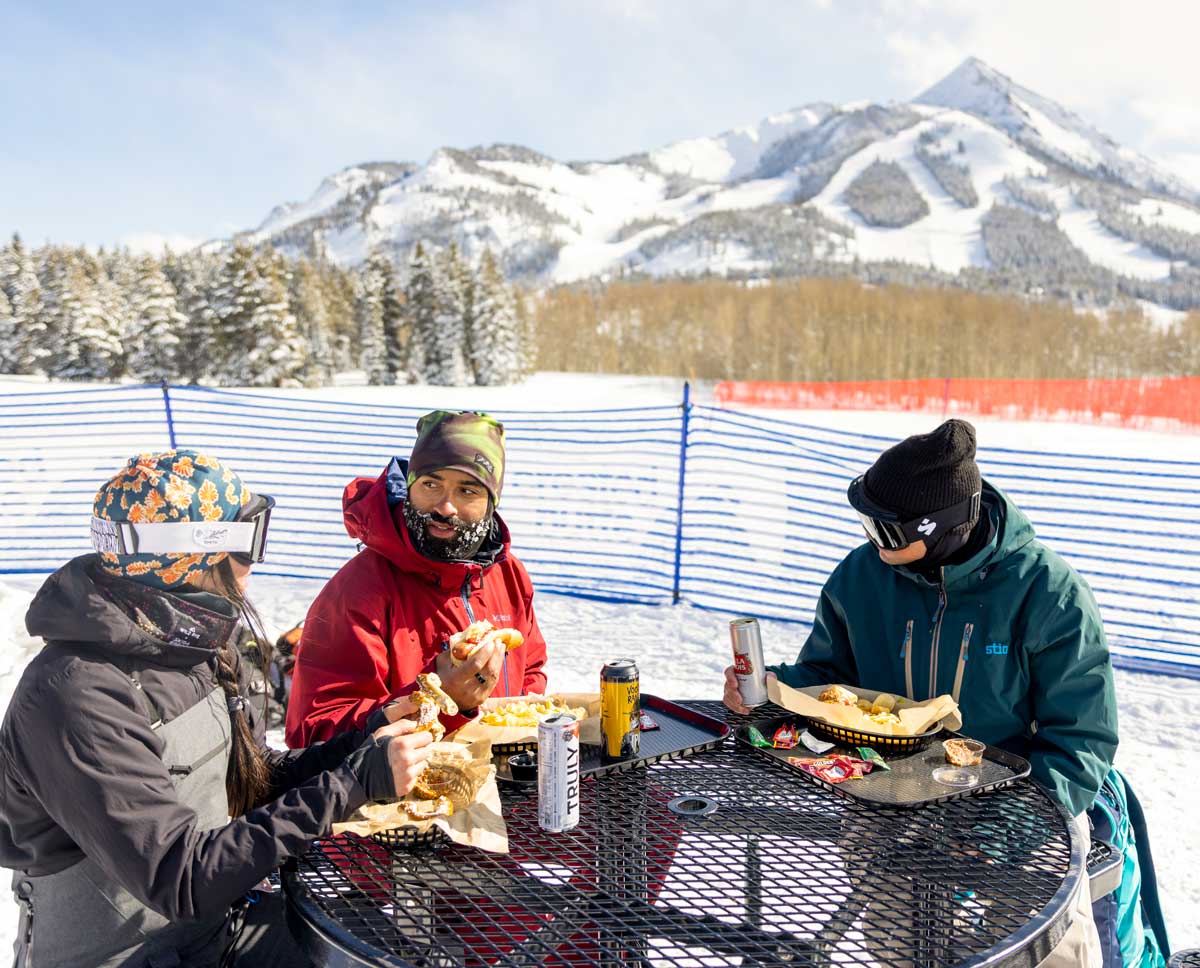 Three people in ski gear eat lunch at a picnic table next to the ski slopes on a sunny day. 