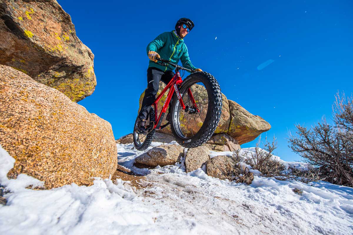 A fat biker and his meaty tires jumps between two rocks onto a snoy trail. The sky is bright blue above him