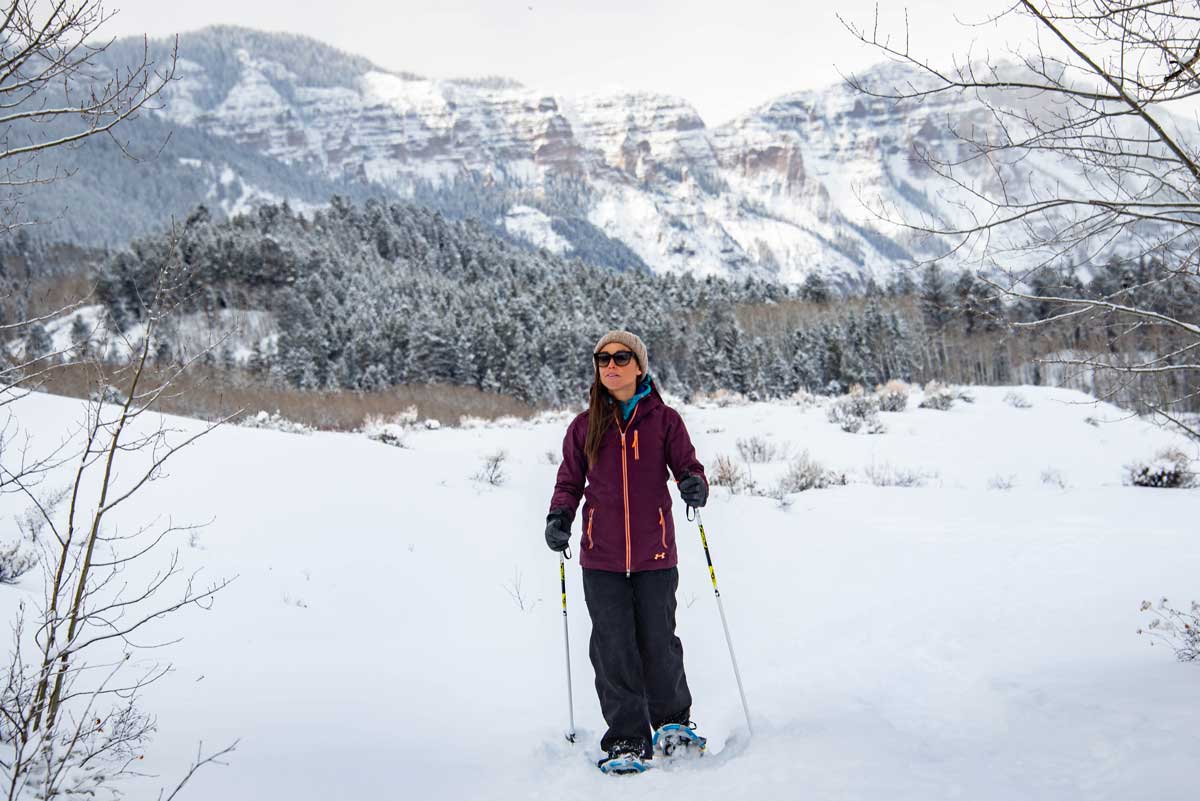 Person snowshoeing on trails outside of Crested Butte. They have poles to help them navigate the powdery trail and snowcapped peaks and evergreens are behind them