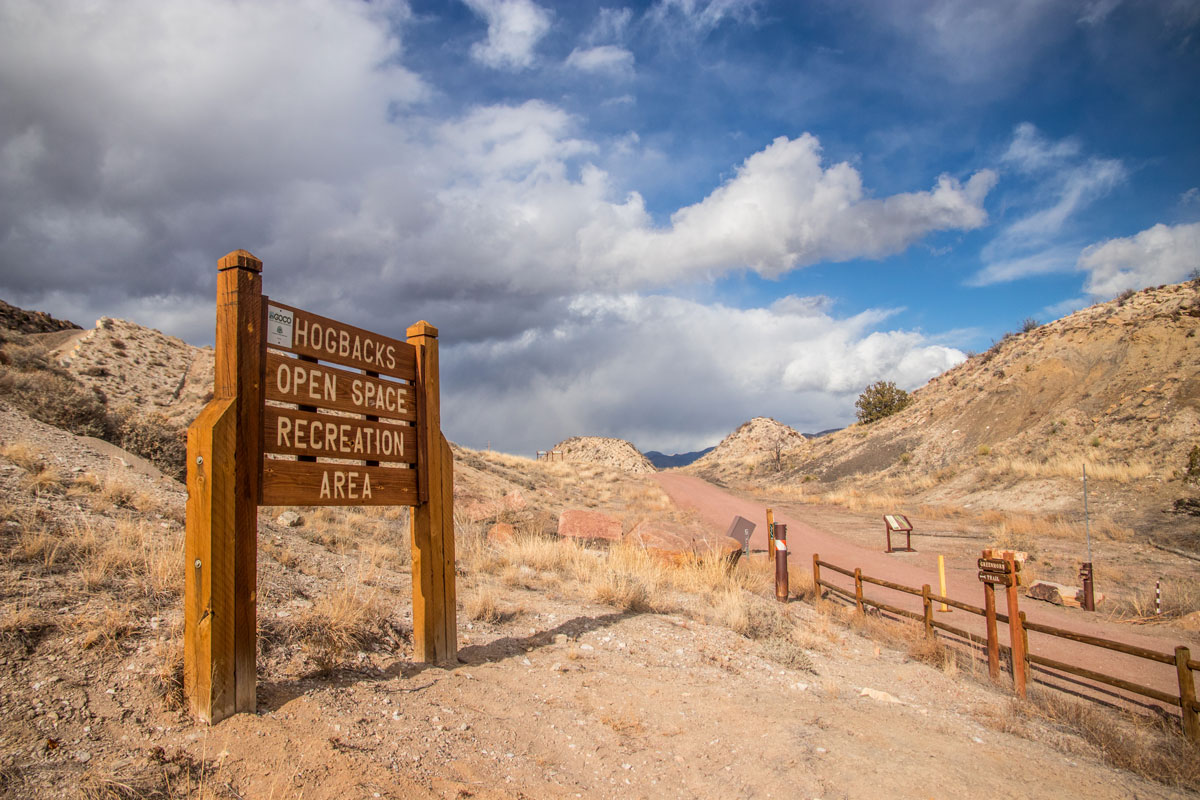 A wooden sign in the foreground labeling a trailhead that says "Hogbacks Open Space Recreation Area." In the background is a cloudy sky above a hiking trail path.