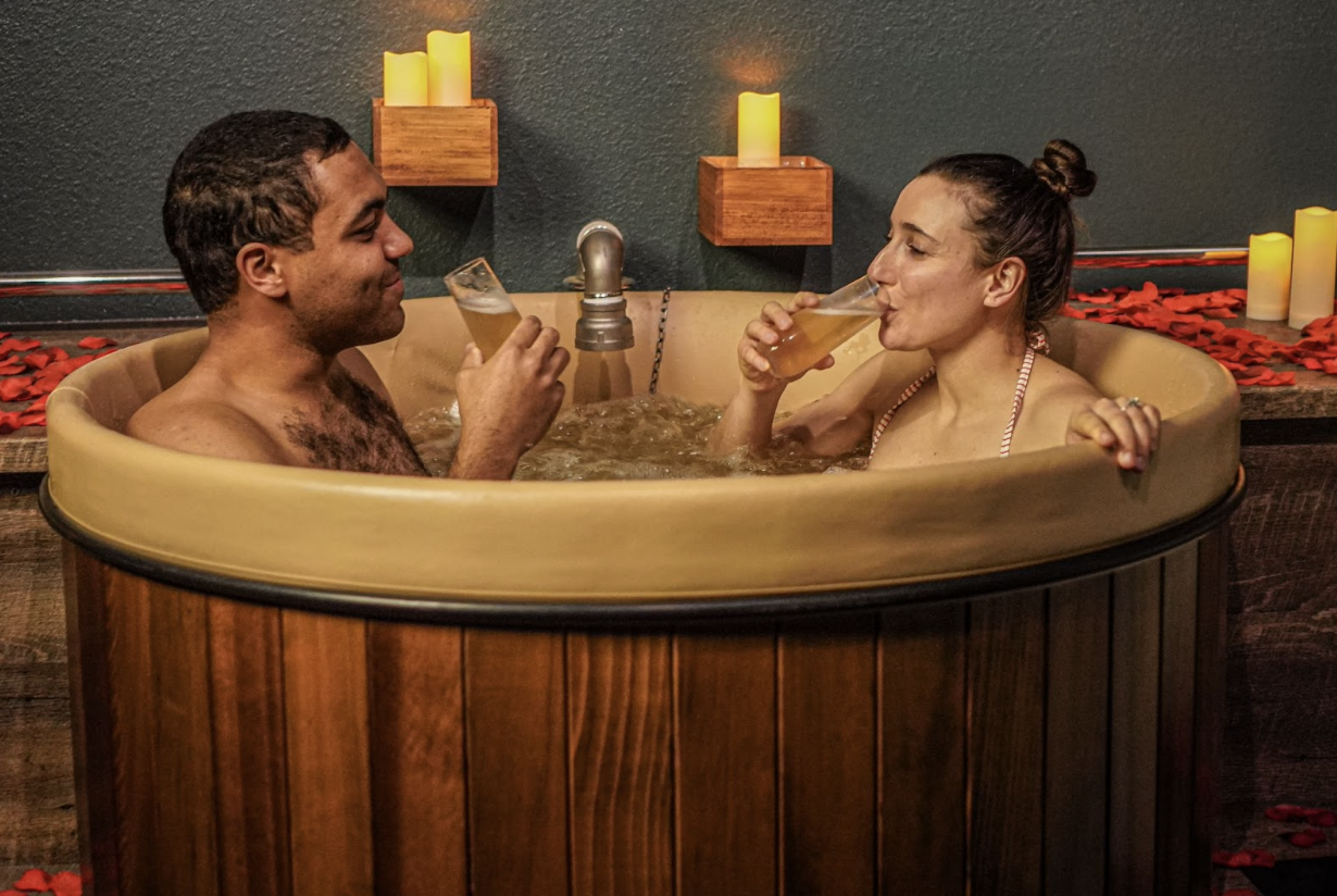 Two people in swimsuits sit in a wooden vat filled with bubbling water and sip tall glasses of pale-yellow beer in Denver, Colorado.