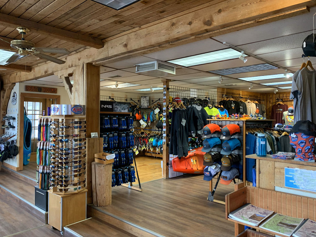 A photo of a retail store adorned in wood paneling on the floors and ceiling. There are a variety of items on shelves, hooks and turntables, including sunglasses, balaclavas, hats, t-shirts, shoes, maps, kayaks, vests, backpacks and more.