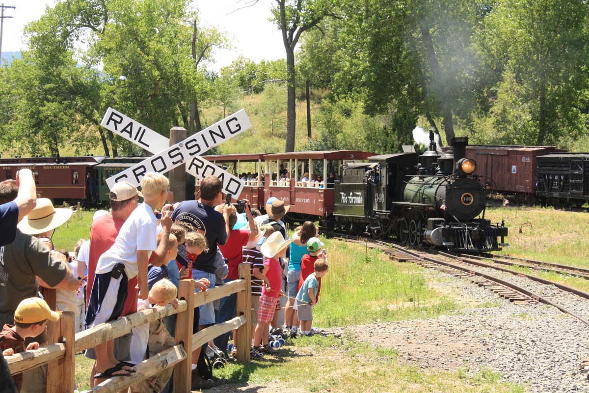A steam train comes around the bend while a group of children and adults watch from behind a fence of a railroad crossing in Colorado.