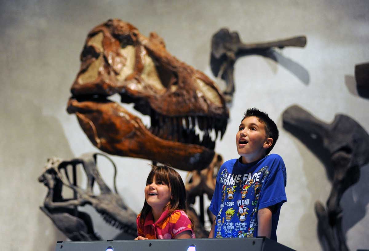 Two kids with giant dinosaur fossils behind them open their mouths in wide wonder at something in front of them we can't see at the Denver Museum of Nature & Science in Colorado.