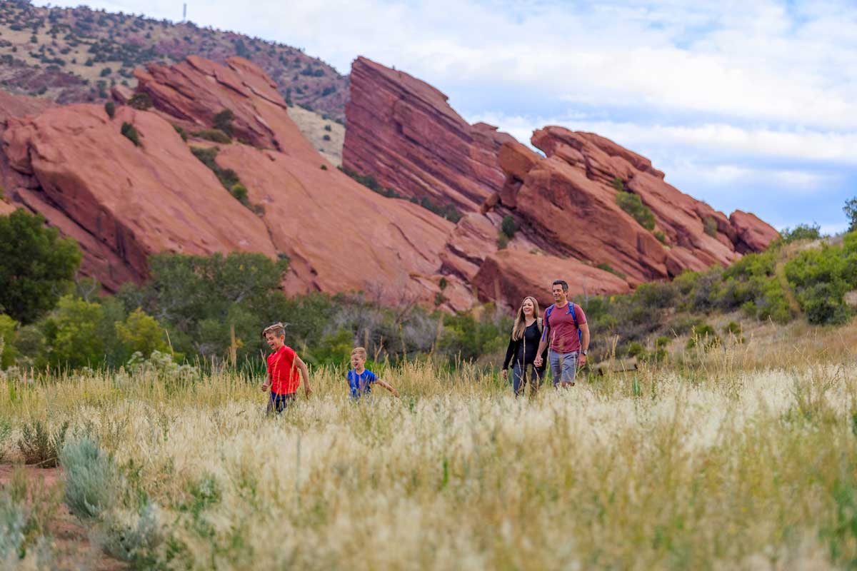 A family of four hikes along a grass-framed trail with giant, sloped slabs of red sandstone reaching into the sky at Red Rocks Park near Denver, Colorado.