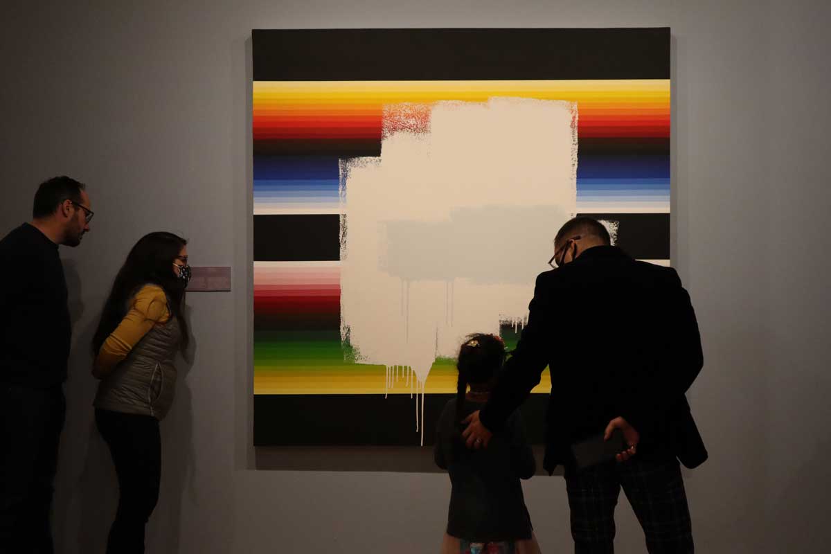 Three adults and a child look at a rainbow-striped painting with a white swatch across the front of it at the Museo de las Americas in Denver, Colorado.