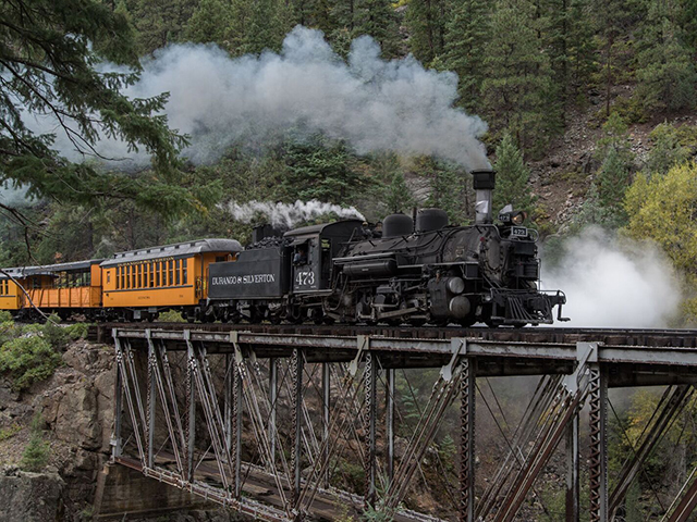 The large Durango Train has steam blowing through its smokestack. The last two visible train cars are painted pumpkin orange with green trim and silver roofs.