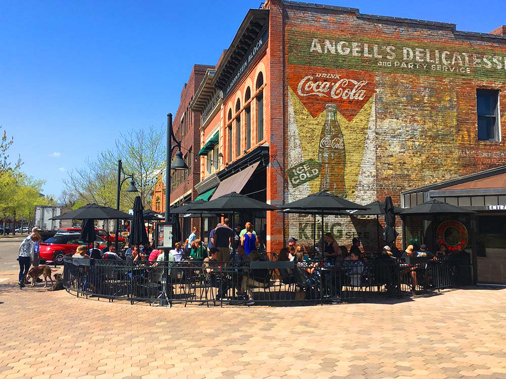 A black-umbrella-covered patio shades diners outside the large, old-looking brick building of CooperSmith's Pub & Brewing restaurant in Fort Collins, Colorado. 