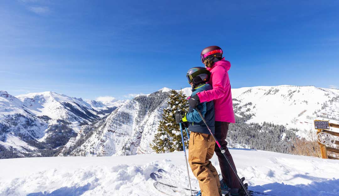 Two skiers stand at the top of a run looking out onto a sunsplashed mountain range