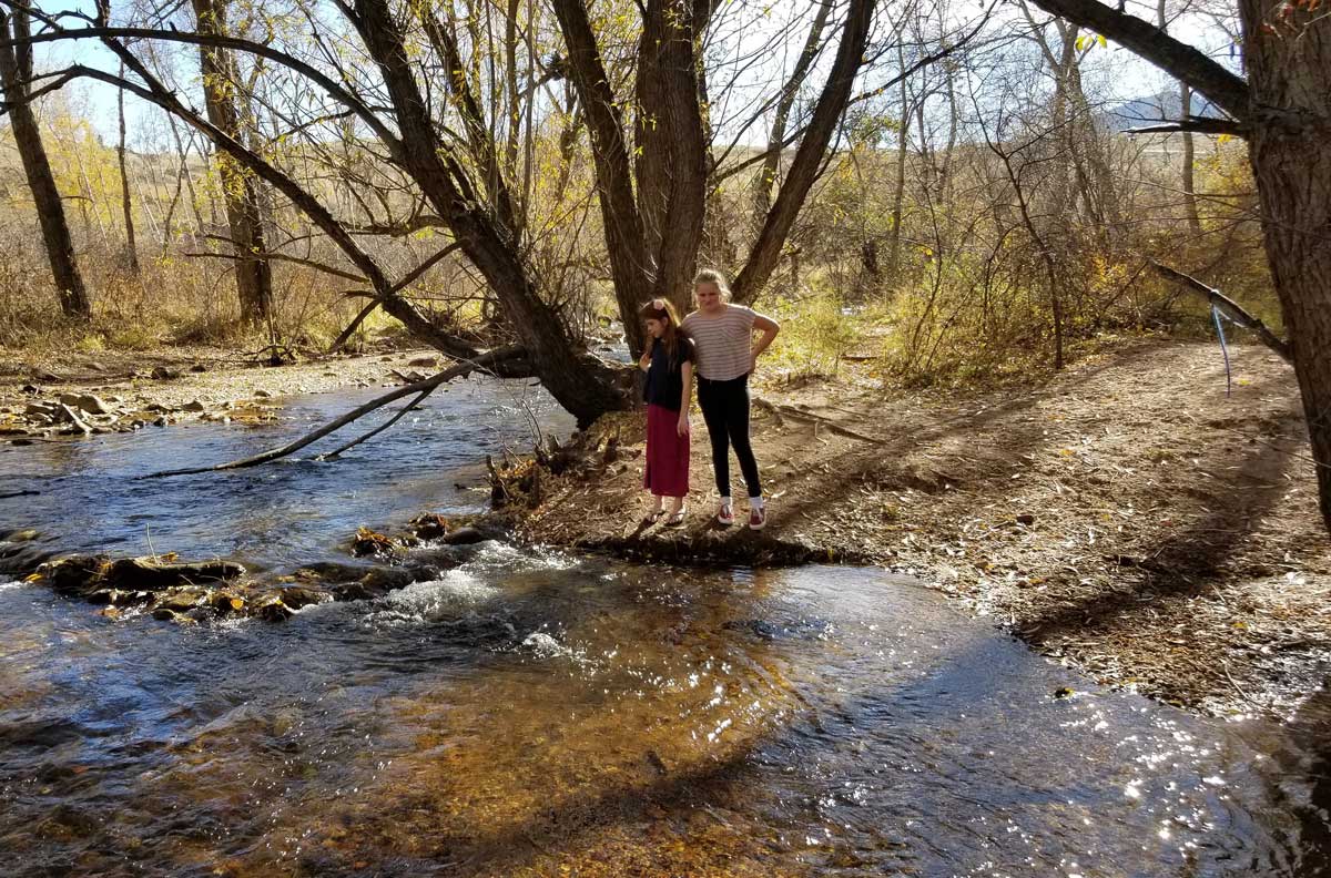 Two girls stand on the banks of a creek next to trees with no leaves