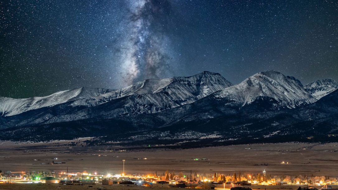 Milky Way and stars above snowy mountains and the lights of a small town at night