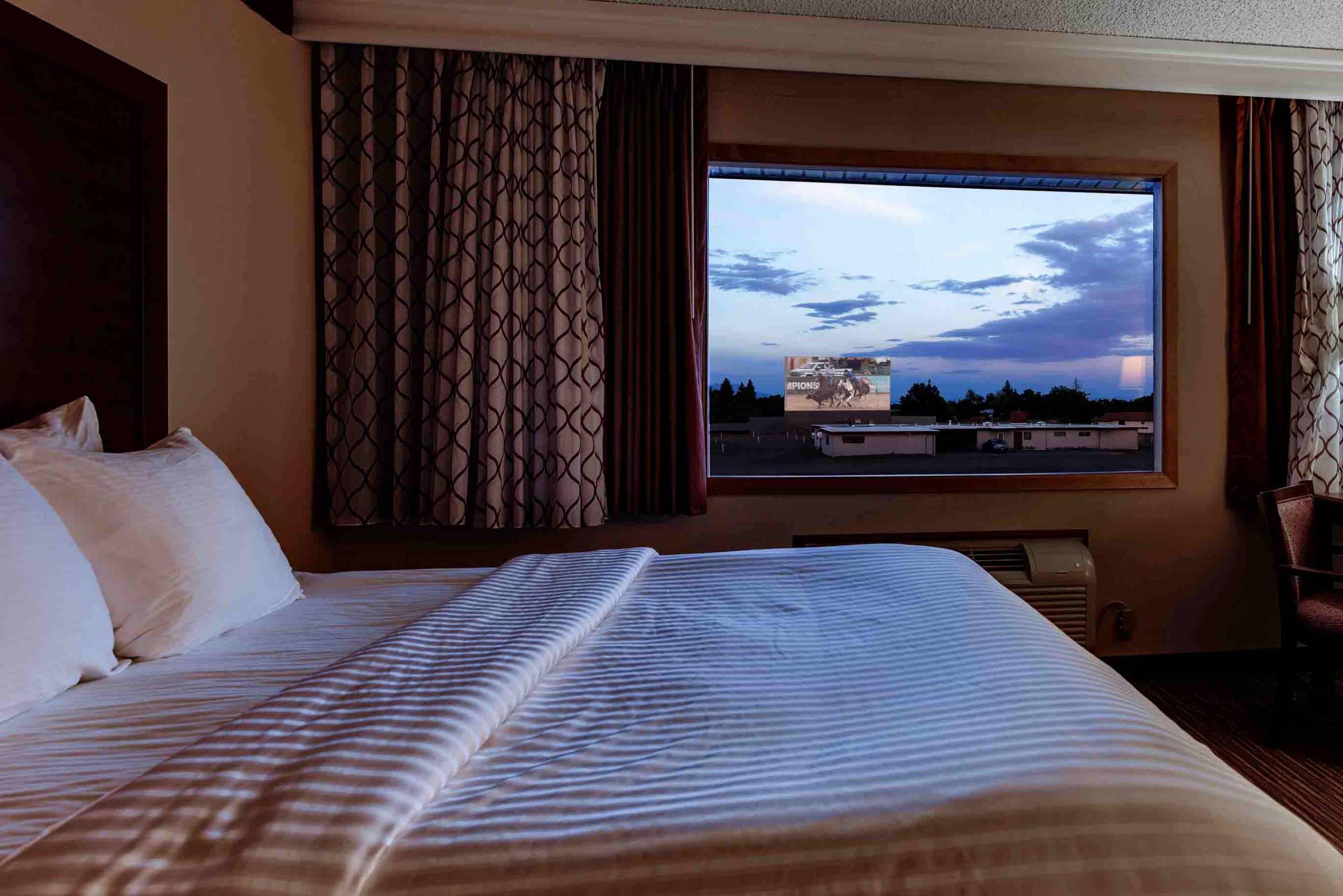 The curtains on a hotel window are pulled open to reveal a drive-in movie screen, visible from the comfort of a cozy, white-blanketed bed at Best Western Movie Manor in Monte Vista Colorado.