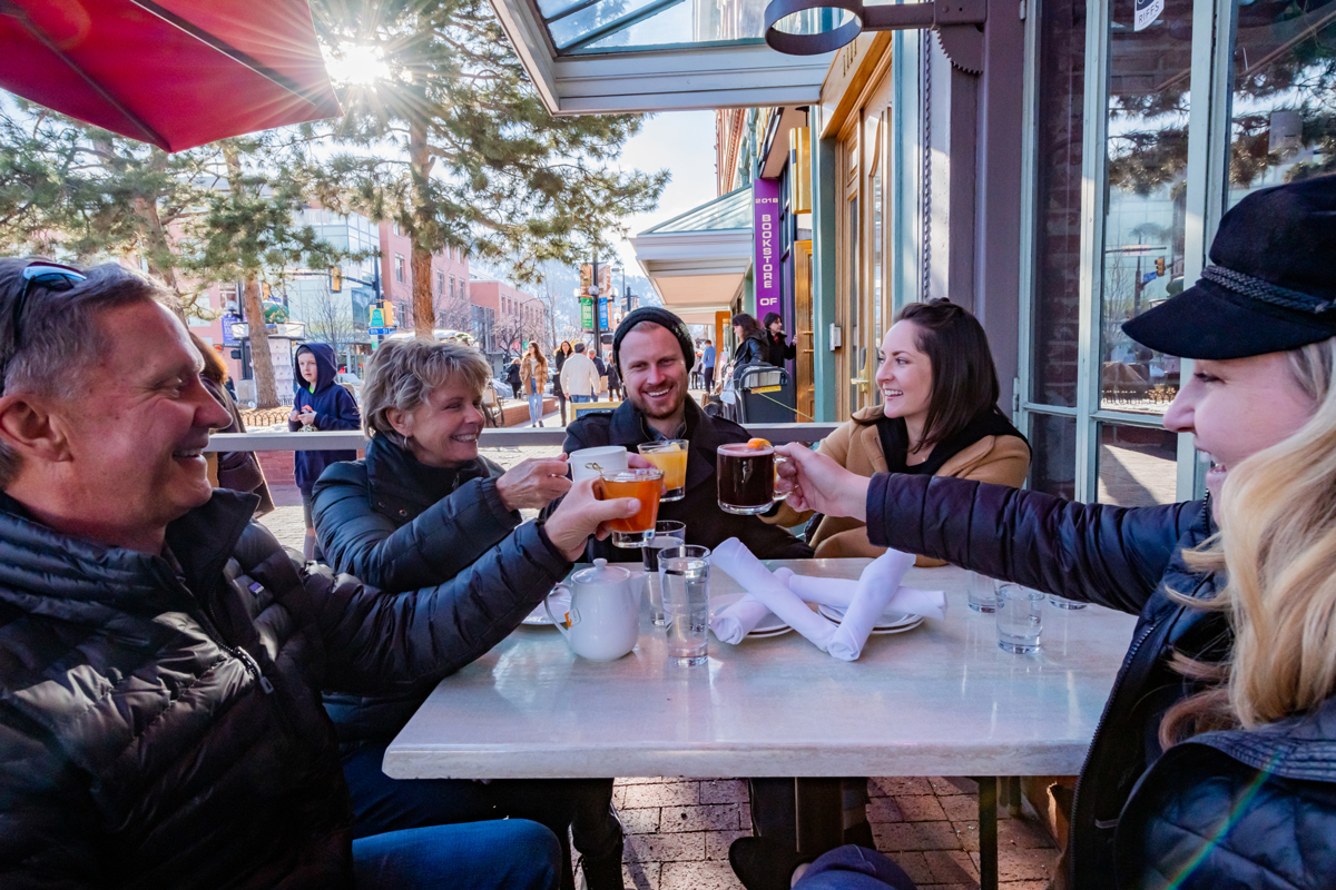 Five members of a family sit around an outdoor table on Pearl Street in Boulder, Colorado, smiling and cheersing their drinks together.