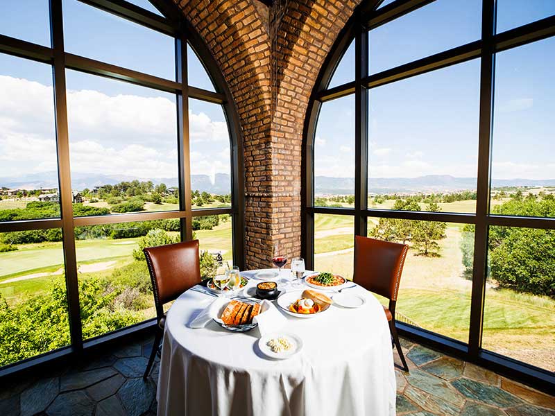 A table with white tablecloth and place settings for two sits in a windowed corner, from which you can see golf courses and mountains
