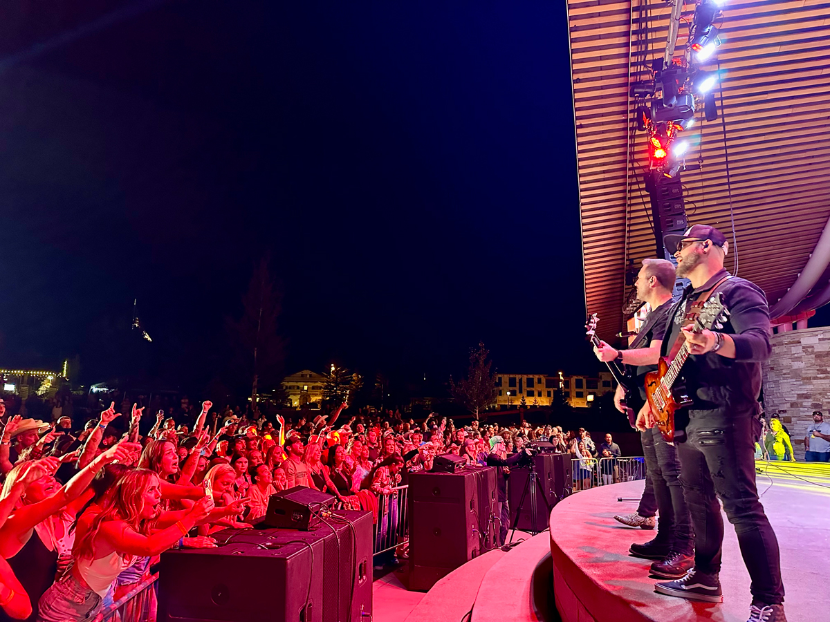 A lively crowd of concert-goers scream and throw their hands in the air as the crowd at a concert in Winter Park. The performers are standing on stage close to the edge, playing guitars.