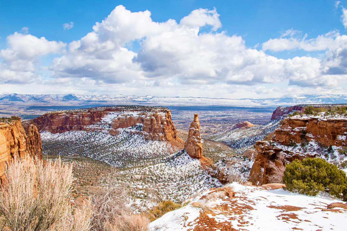 An aerial view of Grand Junction, Colorado in the winter. There is a light dusting of snow on jutting stone formations across the landscape and in the background are more formations and vistas of mountains. The cloud are puffy and gray above the scene.