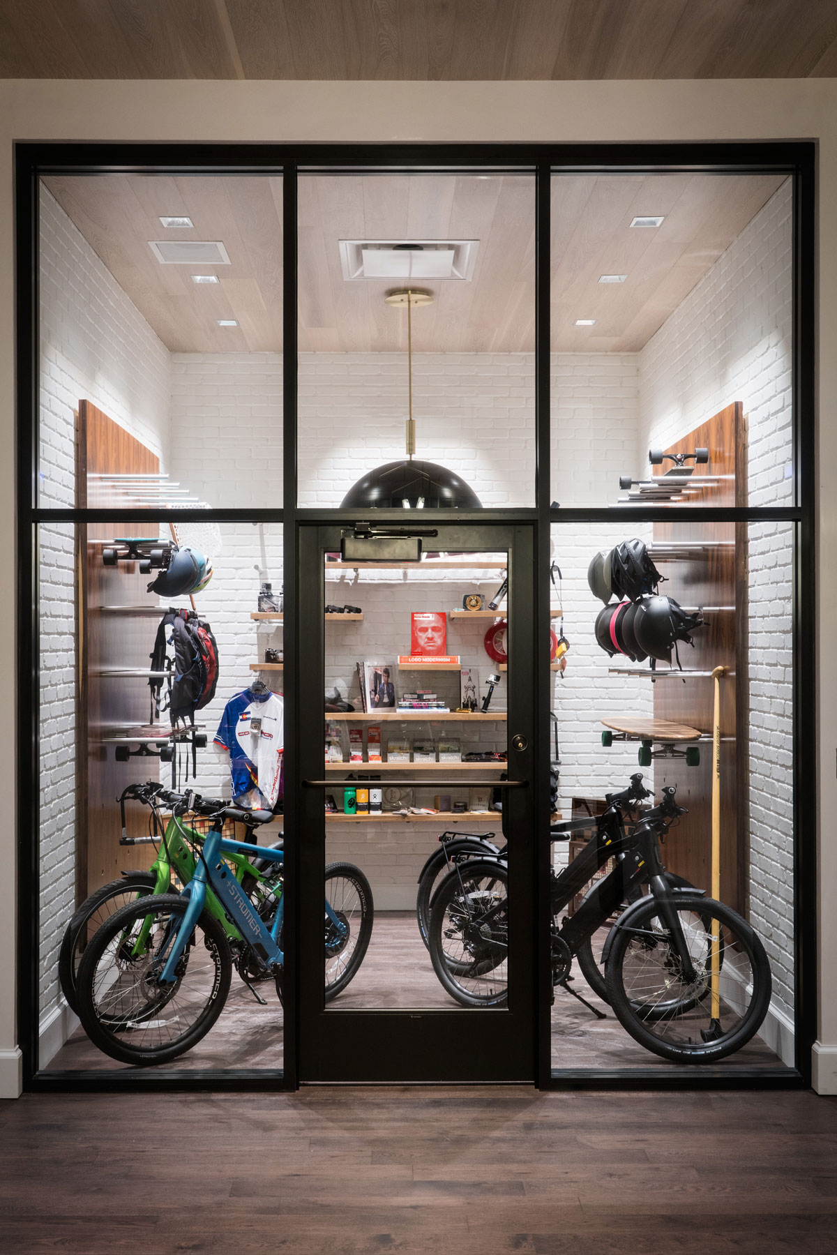 A vertical photo of the Gear Garage at the Halcyon Hotel. There's one glass door surrounded by five wide and tall windows lined with black metal. Inside the door is a small white-brick-walled room with walls of helmets, backpacks, skateboards, bicycles, water bottles, snacks and more.