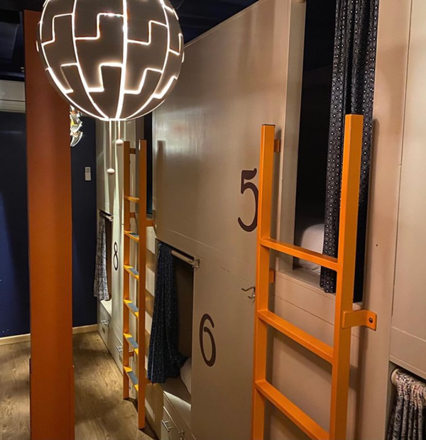 The interior of the Bunkhouse hostel in Minturn, Colorado. On the righthand side of the photo is a beige wall with four rectangular doorways within the wall, two of which have an orange ladder leading up to the doorway, covered in a dark blue curtain. Beyond the curtains are padded white-linen-covered mattresses. Along the sides of each doorway are painted numbers from 5 through 8. Hanging from the ceiling is a geometrical, spherical lighting fixture, whose light carves out little shapes.