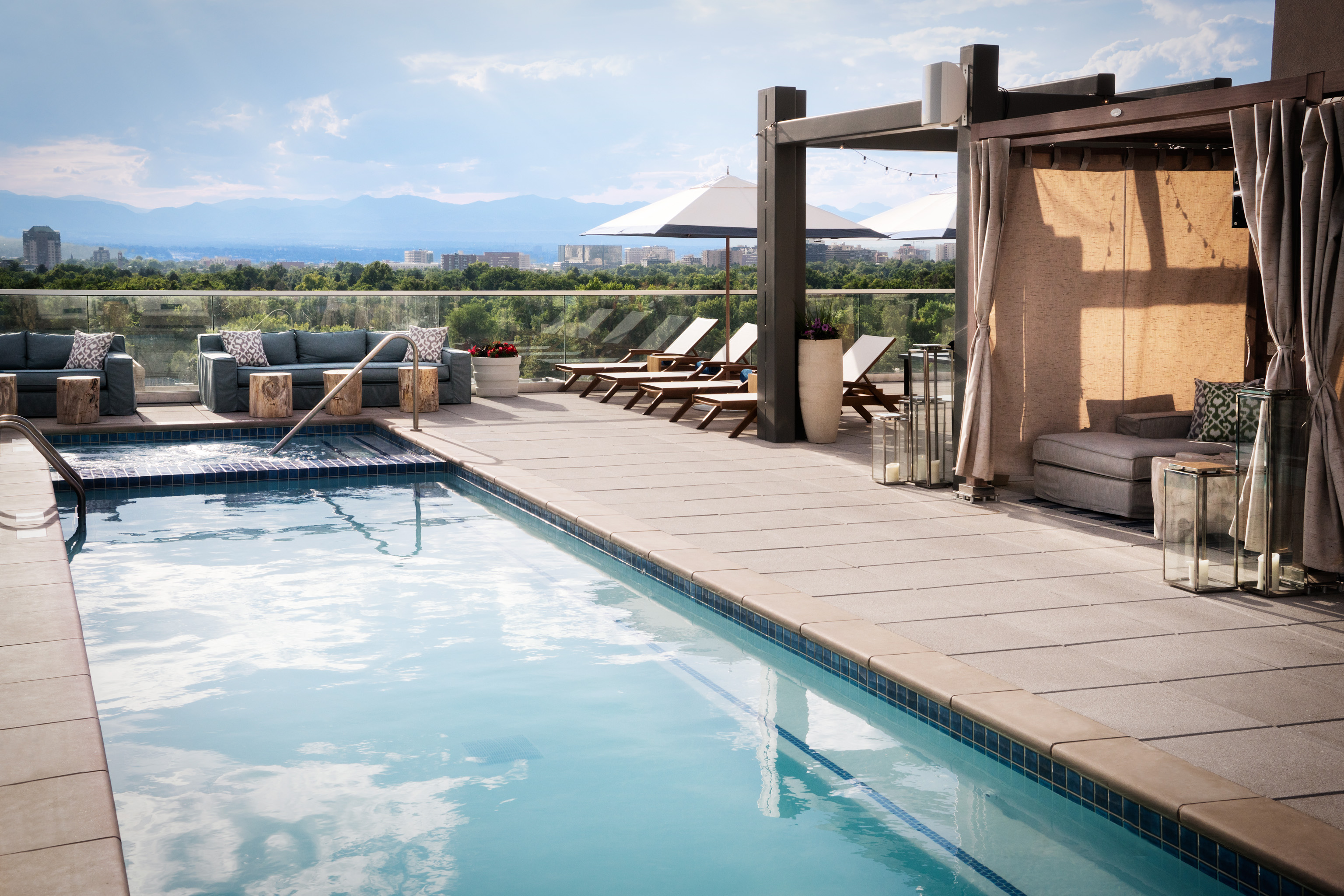 A photo of the long, narrow blue-water pool on the rooftop of the Halcyon hotel in Denver. Around the pool are lounge chairs, draping string lights, awnings and couches. In the distance are groves of green trees and the tall buildings of downtown Denver and the surrounding area. Along the horizon are blue mountain ridges.