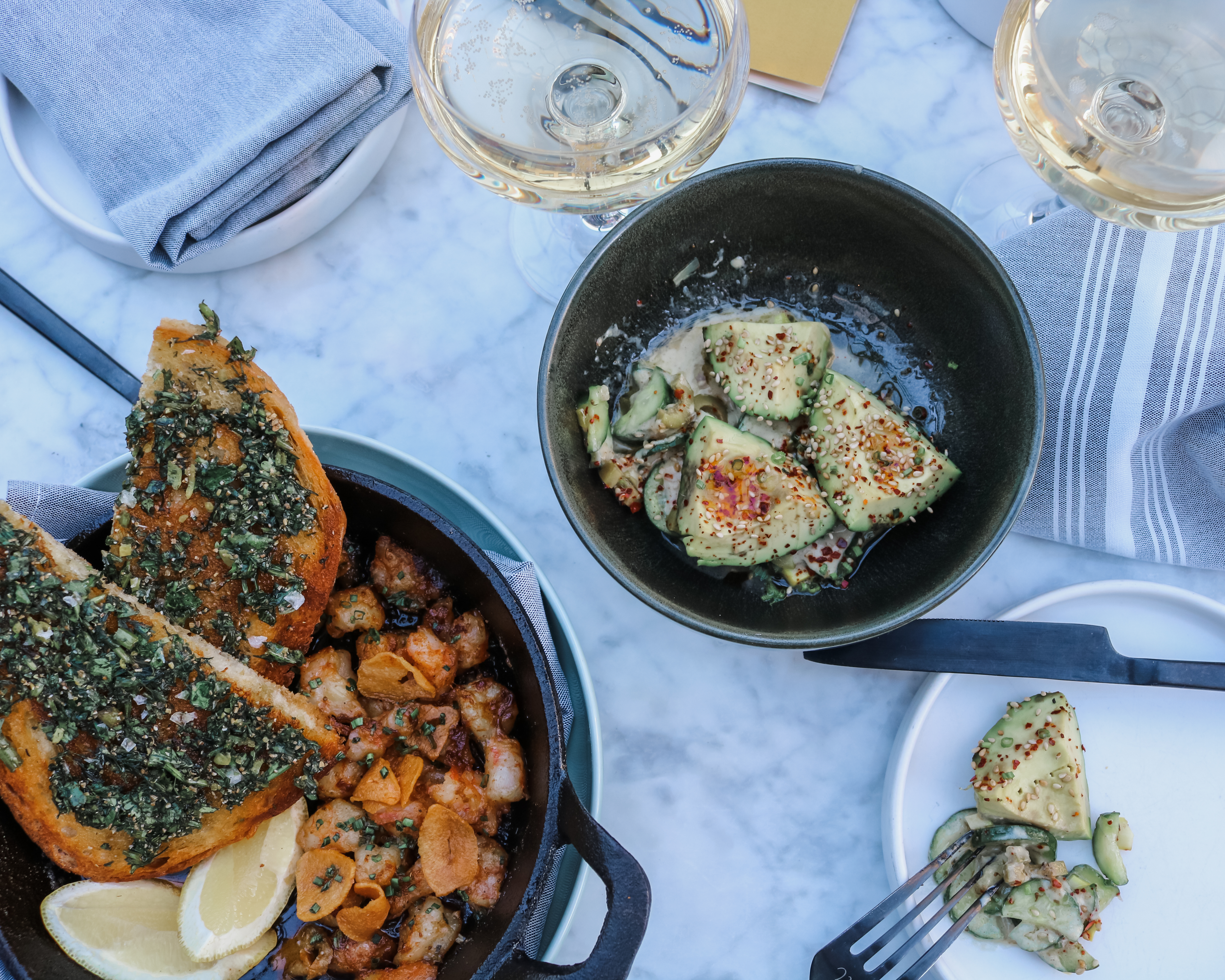 A straight up photo of two dark bowls full of colorful food, including avocados sprinkled in sesame seeds and chili flakes, slices of toast covered in chopped greens and what looks like roasted shrimp and small potatoes. There are glasses of white wine on the nicely set table as well.