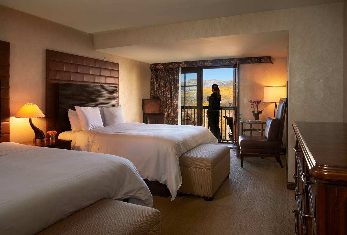 A person with a coffee mug in their hand stands on the patio to survey the view, distant mountains and yellow aspen trees on hills below. The room itself behind the person has two large white-linen-lined beds and there are warm-brown lamps and decorations around the room.