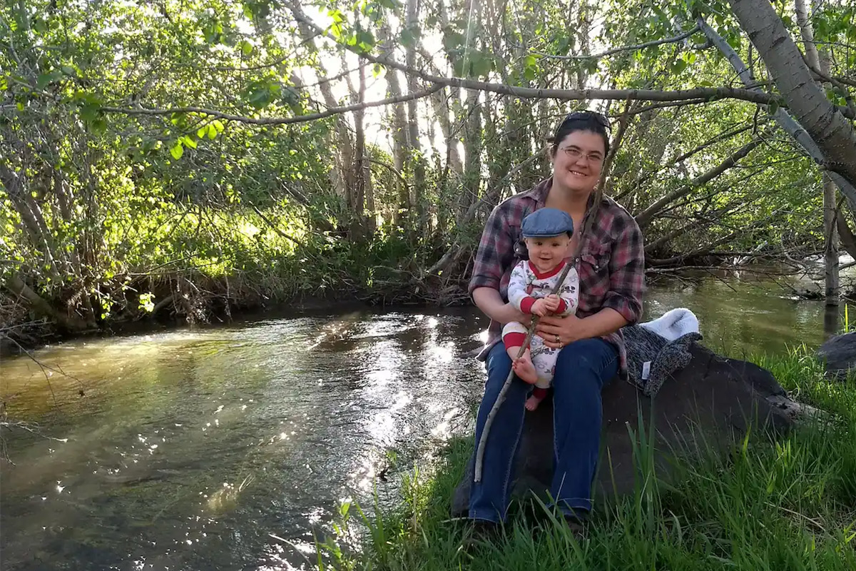 An adult sits on a rock and holds a toddler in their lap near Capulin, Colorado. A babbling creek and lush foliage can be seen in the background.