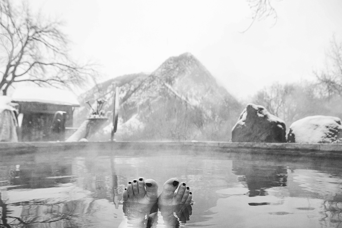 A bather's feet and painted toe nails, stick up through the water of a hot-springs pool at Avalanche Ranch near Redstone, Colorado. A snowy mountain provides a scenic background.