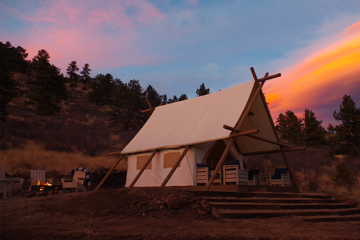 A tent at Black Tree Resort in Colorado stands next to a firepit with chairs. Behind it, the sky is painted with oranges and pinks.