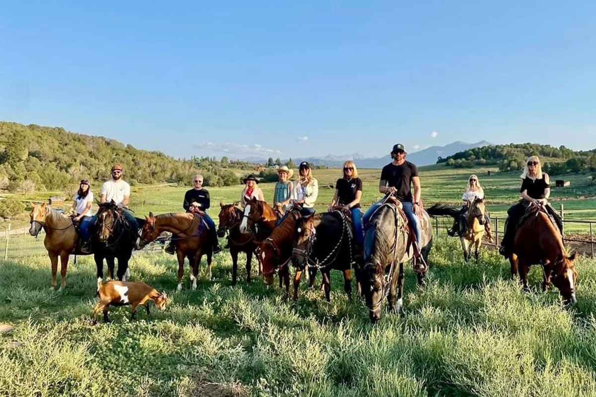 A group of riders at Cedar Ridge Ranch sit atop horses in a rich, green field and pose for the camera. A curious goat wanders into the photo.