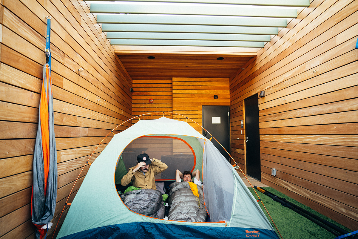 Two glampers stretch in their sleeping bags and prepare to leave their tent. Their tent is set up at the Camp Deck room at Kinship Landing.